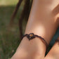 Bijoux de pieds, paire assortie de bracelets de chevilles à ambiance bohème celtique elfique. Fil marron foncé fin tissé en micro macramé avec entrelacs et arabesques, pierre centrale ovale petite taille jaune miel translucide. Bijoux hippie, bohème avec inspiration nature. Chevillères portées sur des chevilles à la peau pâle, bijoux de face 3/4 avec jeu d'ombre et lumière et dreadlocks brunes en fond