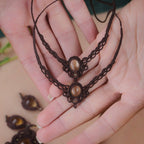 Bijoux de pieds, paire assortie de bracelets de chevilles à ambiance bohème celtique elfique. Fil marron foncé fin tissé en micro macramé avec entrelacs et arabesques, pierre centrale ovale petite taille jaune miel translucide. Bijoux hippie, bohème avec inspiration nature. Chevillères posées à plat dans 2 mains en coupe à la peau pâle, un bijou au dessus de l'autre avec en fond d'autres bijoux marron et jaune miel.