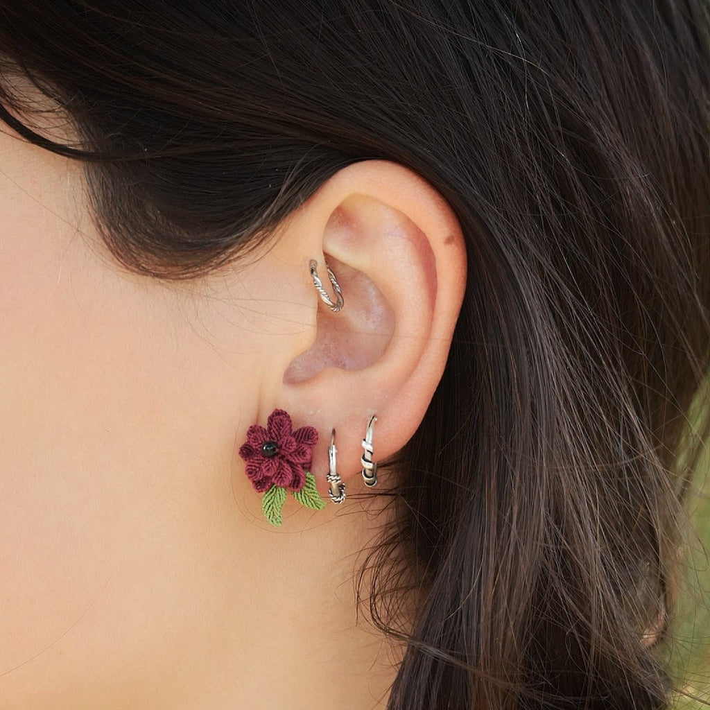 Boucles d’oreilles florales en micro macramé faites main, collection « Pétales éternels ». Bijou artisanal pour femme en coton rouge bordeaux et vert tendre, tissé en fleur 3D avec 13 pétales, 2 feuilles et un cœur en perle d’onyx noire de 3 mm. Fixées sur clous d’oreilles hypoallergéniques en acier inoxydable argenté avec poussoirs papillons. Style bohème, elfique et hippie, idéal pour festivals et costumes féériques. Photo portée sur modèle femme à peau claire et cheveux bruns.