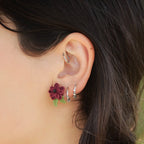 Boucles d’oreilles florales en micro macramé faites main, collection « Pétales éternels ». Bijou artisanal pour femme en coton rouge bordeaux et vert tendre, tissé en fleur 3D avec 13 pétales, 2 feuilles et un cœur en perle d’onyx noire de 3 mm. Fixées sur clous d’oreilles hypoallergéniques en acier inoxydable argenté avec poussoirs papillons. Style bohème, elfique et hippie, idéal pour festivals et costumes féériques. Photo portée sur modèle femme à peau claire et cheveux bruns.