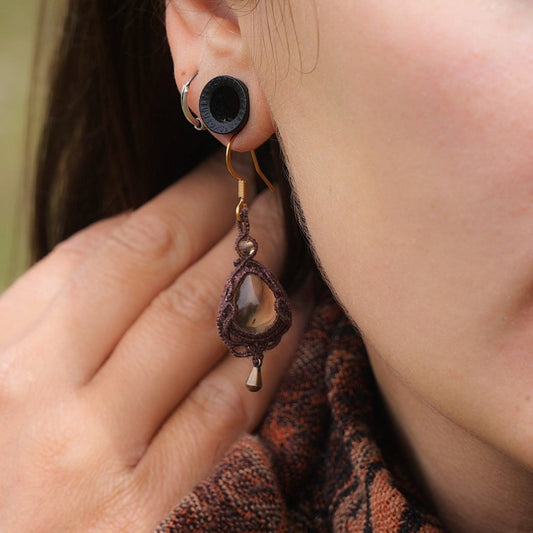 Boucles d'oreilles de style celtique elfique. Bijou féminin composé de : un tissage en relief avec arabesques en micro macramé avec du fil de coton marron foncé chocolat, pierre naturelle de citrine de forme goutte couleur jaune miel accompagné d'une perle ronde en citrine de 3 mm, d'un crochet doré en acier et d'une micro goutte bronze. Le bijou est porté sur une peau dorée et avec un fond de cheveux brun foncé, une main dans le cou avec une écharper aux tons orangers