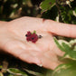 Boucles d’oreilles florales en micro macramé faites main, collection « Pétales éternels », posées sur une main féminine. Fleur 3D tissée en coton rouge bordeaux et vert tendre avec 13 pétales, 2 feuilles et cœur en perle d’onyx noire. Style bohème, elfique et hippie, idéal pour festivals et costumes féériques, photographié au milieu de feuilles et de nature verdoyante.