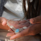 Collier plastron en Larimar Océan bleu ciel, coton couleur lin naturel (beige gris), cauris blancs et perles en acier inoxydable argenté. Système de réglable en tube tissé à l'arrière du bijou. Tissage symétrique en micro macramé avec effet nœuds celtiques entrelacés en relief. Style bohème, hippie, plage, Sirène, tribal et tropical. Convient pour un mariage type bohème ou plage. Bijou tenu vue 3/4, tenue romantique et bohème blanche avec dentelle apparente. Modèle à la peau bronzée et aux cheveux bruns. 