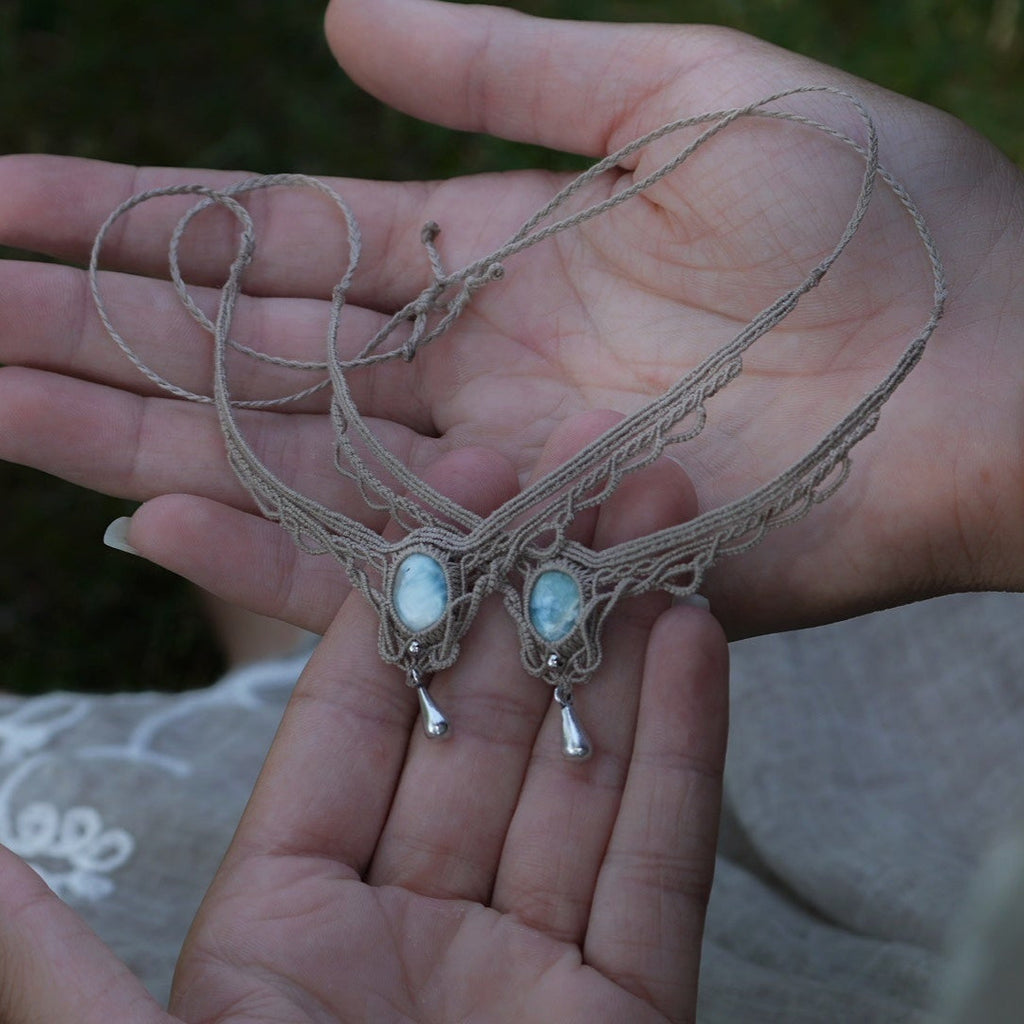 Duo de Bracelets de cheville bohème, ambiance plage et été sur les îles. Bijoux de pieds assortis. Micro macramé en fil de coton couleur lin beige avec une pierre de larimar bleu ciel accompagné d'une perle goutte argenté. Système de réglage à coulisse à l'arrière. Bijoux posé sur 2 mains dans un univers féérique bohème dans un jardin. Modèle avec Dreadlocks brunes, peau caramel et habits assorti au bijou de couleur lin naturel beige gris