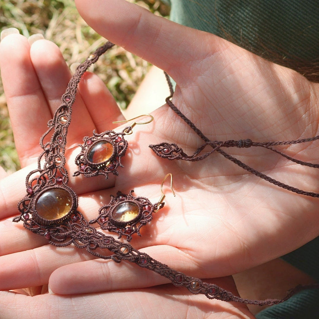 Parure elfique artisanale en micro macramé brun, composée d’un collier ras de cou réglable en citrine jaune et grenat rouge rosé, assorti à des boucles d’oreilles florales. Ensemble bohème et féérique de la collection Clairière de Miel, présenté dans une main féminine à la peau claire.