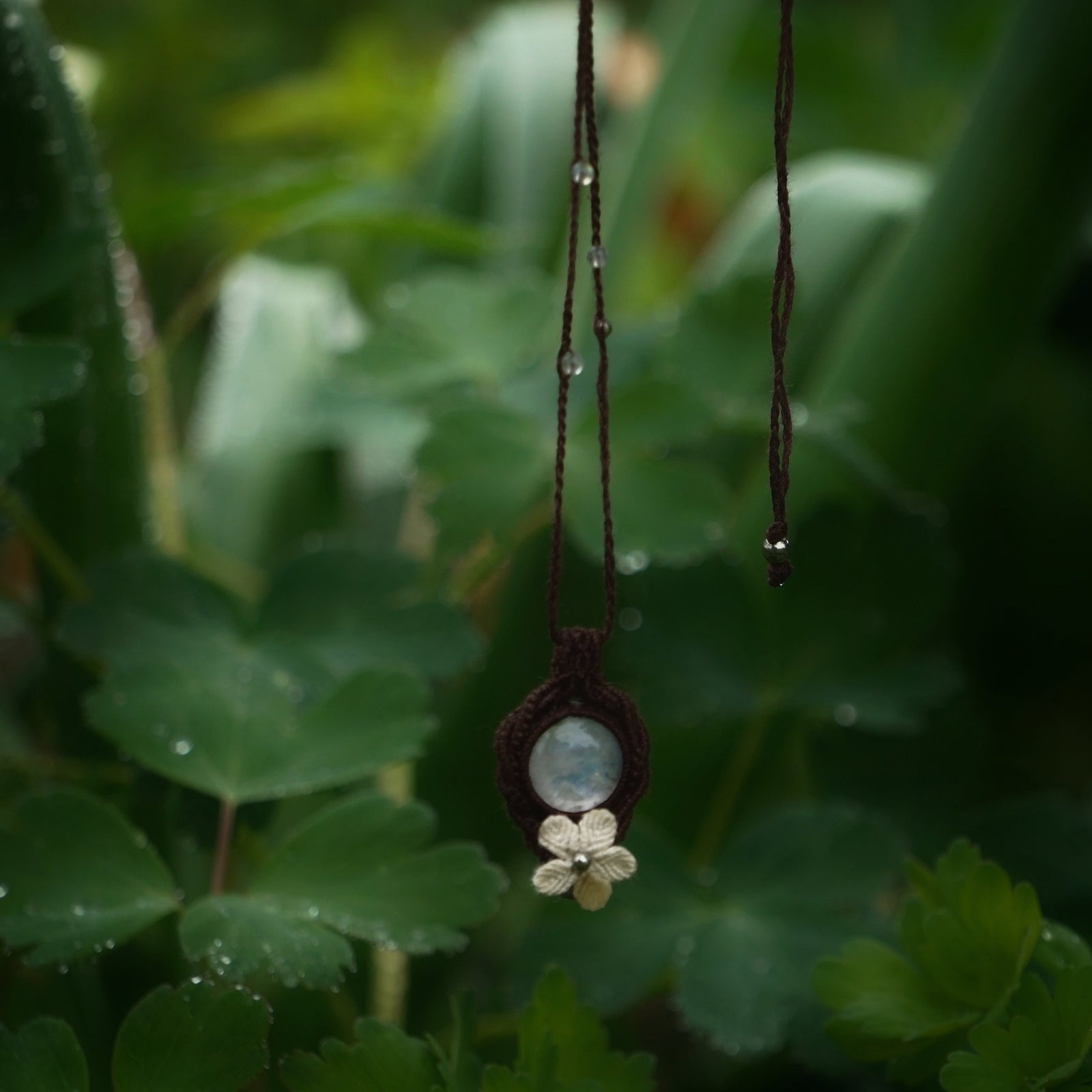 Bijou pendentif en labradorite blanche suspendu verticalement au milieu de de feuilles d'ancolie mouillées par la pluie, collier elfique bohème réglable. Coton et Astrance