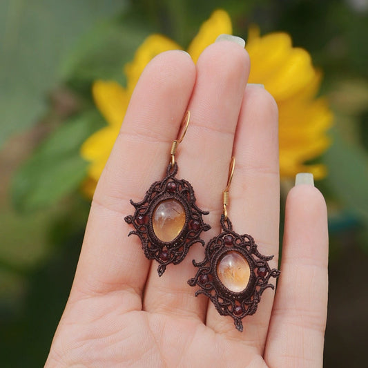 Paire de boucles d'oreilles et d'un collier ras-de-cou elfiques en micro macramé avec pierre naturelle. Bijoux tissé en fil de coton de petit diamètre. Mini Grenats rouge rosé facettés autour d'une citrine jaune ambré, fil de coton marron foncé. Motifs tissés en entrelacs celtiques et arabesques elfiques. Crochet doré. Bijoux ambiance bohème hippie aux couleurs chaudes. Bijoux posés, vue d'en haut dans une main à la peau claire avec tournesol en fond