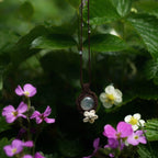 Bijou pendentif en labradorite blanche suspendu verticalement au milieu de fleurs roses et de feuilles de fraisiers mouillées par la pluie, collier elfique bohème réglable.