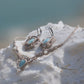 Parure Trésors d'Ecume composée d'une paire de boucles d'oreilles pendentes avec collier ras de cou assorti. Larimar bleu ciel ovales pour les boucles et en poire pointe en bas pour le collier. 3 Perles en nacre blanche pour le collier. Perles acier inoxydable argenté pour les deux pour le côté luxe et précieux. Dormeuses en acier inoxydable pour un port assuré. Fil de Coton couleur lin naturel (beige gris) de 0, 5 mm tissé en micro-macramé en relief version celtique. Bijoux posés sur une pierre blanche 