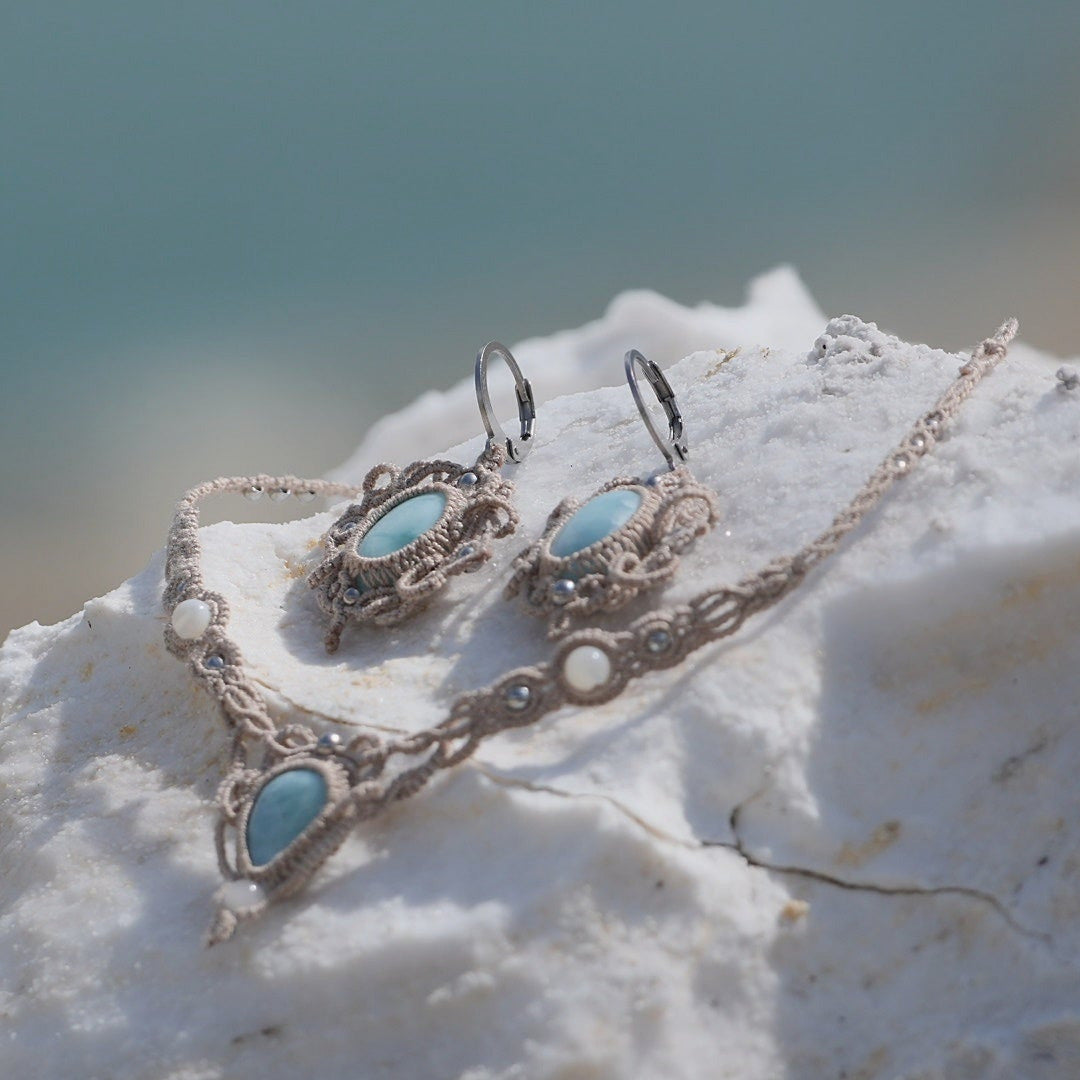 Parure Trésors d'Ecume composée d'une paire de boucles d'oreilles pendentes avec collier ras de cou assorti. Larimar bleu ciel ovales pour les boucles et en poire pointe en bas pour le collier. 3 Perles en nacre blanche pour le collier. Perles acier inoxydable argenté pour les deux pour le côté luxe et précieux. Dormeuses en acier inoxydable pour un port assuré. Fil de Coton couleur lin naturel (beige gris) de 0, 5 mm tissé en micro-macramé en relief version celtique. Bijoux posés sur une pierre blanche 