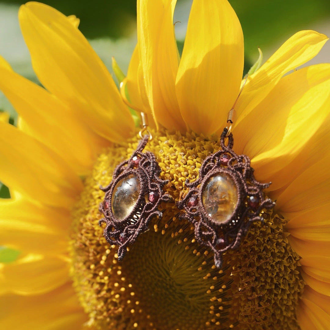 Boucles d'oreilles elfiques en micro macramé avec pierre naturelle. Bijoux tissé en fil de coton de petit diamètre. 4 Mini Grenats rouge rosé facettés autour d'une citrine jaune ambré, fil de coton marron foncé. Motifs tissés en entrelacs celtiques et arabesques elfiques. Crochet doré. Bijou ambiance bohème hippie aux couleurs chaudes. Bijou posé à la verticale, vue de 3/4 sur un tournesol jaune 