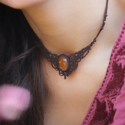Collier ras-de-cou elfiques en micro macramé avec pierre naturelle. Bijoux tissé en fil de coton de petit diamètre. Mini Grenats rouge rosé facettés autour d'une citrine jaune ambré ovale, fil de coton marron foncé. Motifs tissés en entrelacs celtiques et arabesques elfiques Forme V. Bijoux ambiance bohème hippie aux couleurs chaudes. Bijoux porté sur un mannequin féminin à la eau claire et cheveux bruns, habit rose fuchsia. Vue de 3/4