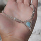 Collier ras-de-cou forme v Trésors d'Ecume composée d'une Larimar bleu ciel en poire pointe en bas, perles en nacre blanche ronde rappel des coquillages et de la plage, mini Perles rondes en acier inoxydable argenté pour le côté luxe et précieux. Fil de Coton couleur lin naturel (beige gris) de 0, 5 mm tissé en micro-macramé en relief version celtique. Collage 3 photos : Bijoux à plat dans une main à la peau claire. Rocher blanc en fond. . Photo ambiance tropiques et plages, style bohème hippie 
