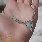 Collier ras-de-cou forme v Trésors d'Ecume composée d'une Larimar bleu ciel en poire pointe en bas, perles en nacre blanche ronde rappel des coquillages et de la plage, mini Perles rondes en acier inoxydable argenté pour le côté luxe et précieux. Fil de Coton couleur lin naturel (beige gris) de 0, 5 mm tissé en micro-macramé en relief version celtique. Collage 3 photos : Bijoux à plat dans une main à la peau claire. Rocher blanc en fond. . Photo ambiance tropiques et plages, style bohème hippie 