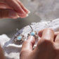 Boucles d'oreilles Trésors d'Ecume composée d'une Larimar bleu ciel ovales, mini Perles rondes en acier inoxydable argenté pour le côté luxe et précieux. Dormeuses en acier inoxydable pour un port assuré. Fil de Coton couleur lin naturel (beige gris) de 0, 5 mm tissé en micro-macramé en relief version celtique. Collage 3 photos : Bijoux posés et porté sur une femme bronzée et brune, et descriptif de composition visuel avec flèche 