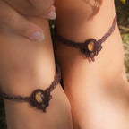 Bijoux de pieds, paire assortie de bracelets de chevilles à ambiance bohème celtique elfique. Fil marron foncé fin tissé en micro macramé avec entrelacs et arabesques, pierre centrale ovale petite taille jaune miel translucide. Bijoux hippie, bohème avec inspiration nature. Chevillères portées sur des chevilles à la peau pâle, bijoux de face avec jeu d'ombre et lumière et fleurs jaune en haut à droite.