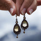 Boucles d'oreilles de style celtique elfique. Bijou féminin composé de : un tissage en relief avec arabesques en micro macramé avec du fil de coton marron foncé chocolat, pierre naturelle de citrine de forme goutte couleur jaune miel accompagné d'une perle ronde en citrine de 3 mm, d'un crochet doré en acier et d'une micro goutte bronze. Le bijou est présenté de façon verticale avec un fond montagneux bleuté