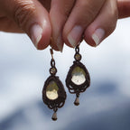 Boucles d'oreilles de style celtique elfique. Bijou féminin composé de : un tissage en relief avec arabesques en micro macramé avec du fil de coton marron foncé chocolat, pierre naturelle de citrine de forme goutte couleur jaune miel accompagné d'une perle ronde en citrine de 3 mm, d'un crochet doré en acier et d'une micro goutte bronze. Le bijou est présenté de façon verticale avec un fond montagneux bleuté