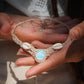 Collier plastron en Larimar Océan bleu ciel, coton couleur lin naturel (beige gris), cauris blancs et perles en acier inoxydable argenté. Système de réglable en tube tissé à l'arrière du bijou. Tissage symétrique en micro macramé avec effet nœuds celtiques entrelacés en relief. Style bohème, hippie, plage, Sirène, tribal et tropical. Convient pour un mariage type bohème ou plage. Bijou tenu vue 3/4, tenue romantique et bohème blanche avec dentelle apparente. Modèle à la peau bronzée et aux cheveux bruns. 