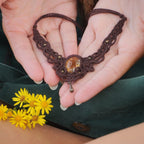 Collier large en micro macramé de couleur marron foncé, jaune ambré et bronze. Motif tissé en micro macramé avec ambiance entrelacs celtiques et arabesques elfiques. Présence d'une pierre centrale ovale de grande taille jaune translucide, gouttes bronze, forme V avec mini perles ronde bronze parsemées. Photo ambiance bohème hippie avec une touche d'élégance. Bijou posé à plat dans 2 mains en coupe à peau claire. Fleur jaune en premier plan et jupe vert forêt en velours côtelé à l'arrière