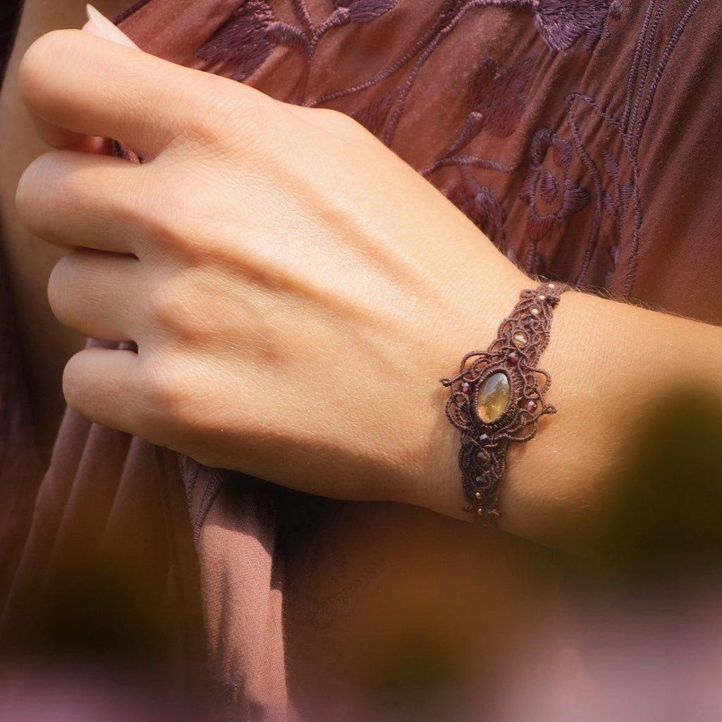 Bracelet large elfique en micro macramé brun, orné d’une citrine jaune pâle et de perles de grenat rouge rosé. Bijou bohème et artisanal réglable, porté au poignet d’une femme à la peau claire.