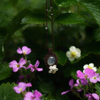 Bijou pendentif en labradorite blanche suspendu verticalement au milieu de fleurs roses et de feuilles de fraisiers mouillées par la pluie, collier elfique bohème réglable. Coton et Astrance