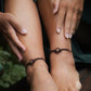Bijoux de pieds, paire assortie de bracelets de chevilles à ambiance bohème celtique elfique. Fil marron foncé fin tissé en micro macramé avec entrelacs et arabesques, pierre centrale ovale petite taille jaune miel translucide. Bijoux hippie, bohème avec inspiration nature. Chevillères portées sur des chevilles à la peau pâle, ambiance sombre/dark. Modèle assise sur un banc en bois, les jambes relevées, les mains autour des mollets, de face avec jeu d'ombre en fond, jupe velours côtelé vert sapin