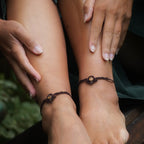 Bijoux de pieds, paire assortie de bracelets de chevilles à ambiance bohème celtique elfique. Fil marron foncé fin tissé en micro macramé avec entrelacs et arabesques, pierre centrale ovale petite taille jaune miel translucide. Bijoux hippie, bohème avec inspiration nature. Chevillères portées sur des chevilles à la peau pâle, ambiance sombre/dark. Modèle assise sur un banc en bois, les jambes relevées, les mains autour des mollets, de face avec jeu d'ombre en fond, jupe velours côtelé vert sapin