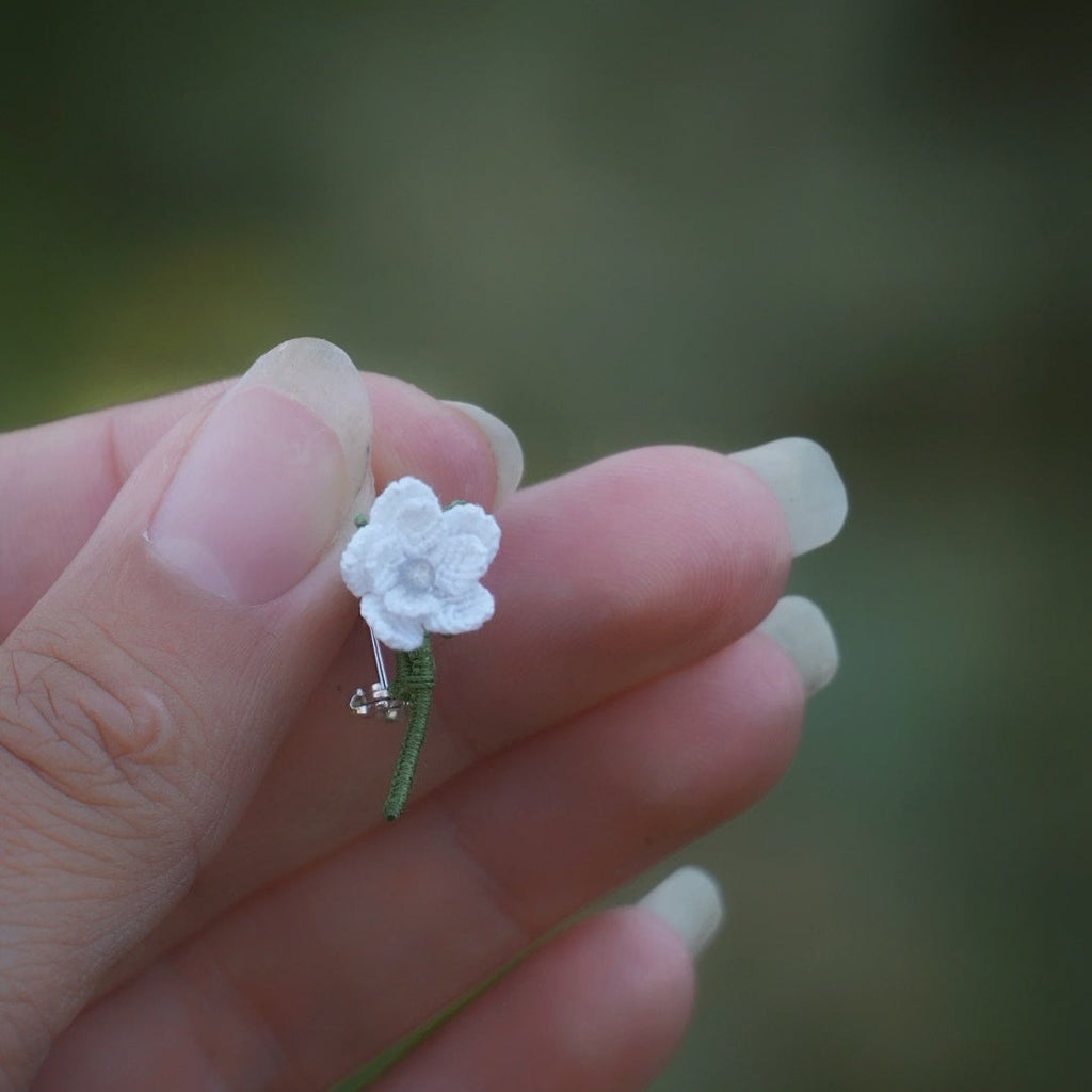 Broche miniature florale blanche en micro macramé avec labradorite bleu-vert au centre, tenue dans une main sur fond de feuilles vertes, bijou bohème et elfique artisanal.