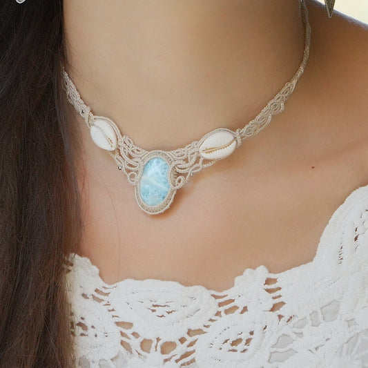 Collier plastron en Larimar Océan bleu ciel, coton couleur lin naturel (beige gris), cauris blancs et perles en acier inoxydable argenté. Système de réglable en tube tissé à l'arrière du bijou. Tissage symétrique en micro macramé avec effet nœuds celtiques entrelacés en relief. Style bohème, hippie, plage, Sirène, tribal et tropical. Convient pour un mariage type bohème ou plage. Bijou porté avec une tenue romantique et bohème blanche avec dentelle apparente. Modèle à la peau bronzée et aux cheveux bruns. 