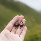 Boucles d'oreilles de style celtique elfique. Bijou féminin composé de : un tissage en relief avec arabesques en micro macramé avec du fil de coton marron foncé chocolat, pierre naturelle de citrine de forme goutte couleur jaune miel accompagné d'une perle ronde en citrine de 3 mm, d'un crochet doré en acier et d'une micro goutte bronze. Le bijou est posé dans une main pâle avec un fond nature vert