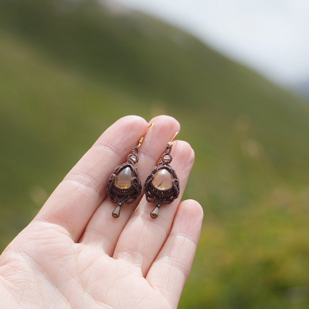 Boucles d'oreilles de style celtique elfique. Bijou féminin composé de : un tissage en relief avec arabesques en micro macramé avec du fil de coton marron foncé chocolat, pierre naturelle de citrine de forme goutte couleur jaune miel accompagné d'une perle ronde en citrine de 3 mm, d'un crochet doré en acier et d'une micro goutte bronze. Le bijou est posé dans une main pâle avec un fond nature vert