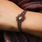 Bracelet large elfique en micro macramé brun, orné d’une citrine jaune pâle et de perles de grenat rouge rosé. Bijou bohème et artisanal réglable, porté au poignet d’une femme à la peau claire.
