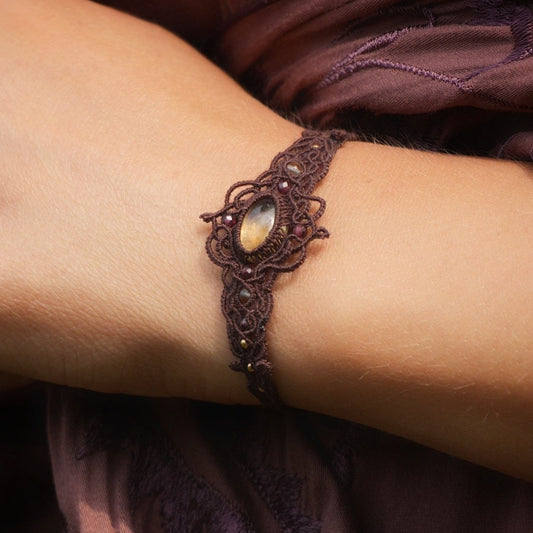 Bracelet réglable en micro macramé. Bijou féminin composé d'une citrine jaune ovale translucide au centre, entourée de 6 perles en grenat rouge à facettes de 3 mm puis de 3 perles dorées de 2 mm. Fil de coton de 0.4 mm tissé en micro macramé avec de motifs en reliefs type nœuds celtiques et motifs arabesques  féériques et elfiques. Bijou large ambiance et style bohème hippie, fairycore.

Bijou porté sur un poignet de femme  la peau claire, cheveux marron, prise de vue de face et vêtement parme brodé floral.