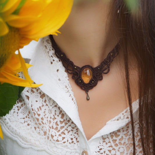 Collier large en micro macramé de couleur marron foncé, jaune ambré et bronze. Motif tissé en micro macramé avec ambiance entrelacs celtiques et arabesques elfiques. Présence d'une pierre centrale ovale de grande taille jaune translucide, gouttes bronze, forme V avec mini perles ronde bronze parsemées. Photo ambiance bohème hippie avec une touche d'élégance. Bijou porté sur une peau claire avec tournesol jaune en premier plan et chemise blanche en dentelle.