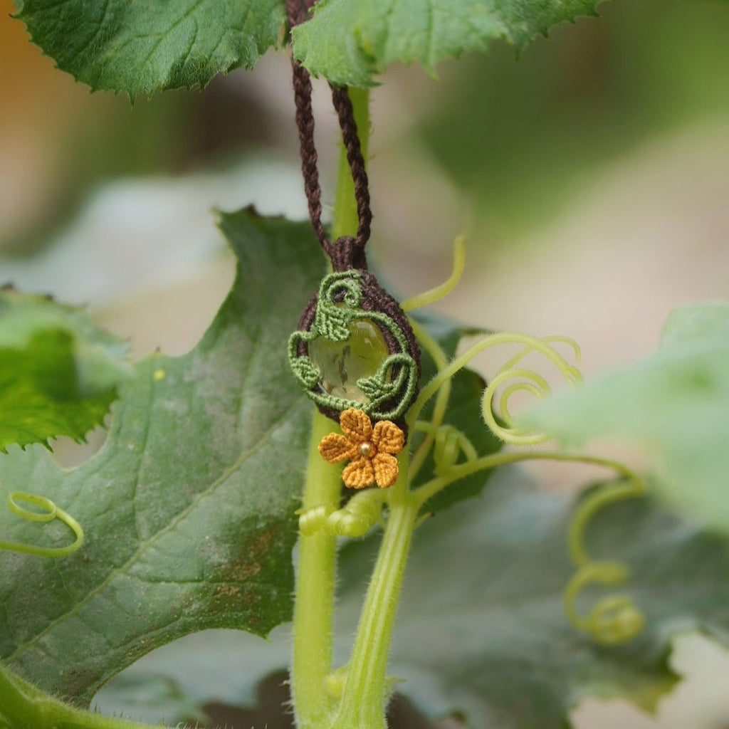 Collier elfique floral en citrine et macramé, fait main en France. Pièce artisanale réglable de la collection Clairière de Miel, photographiée au cœur de la nature parmi des feuilles de courge pour souligner son inspiration bohème et féérique.