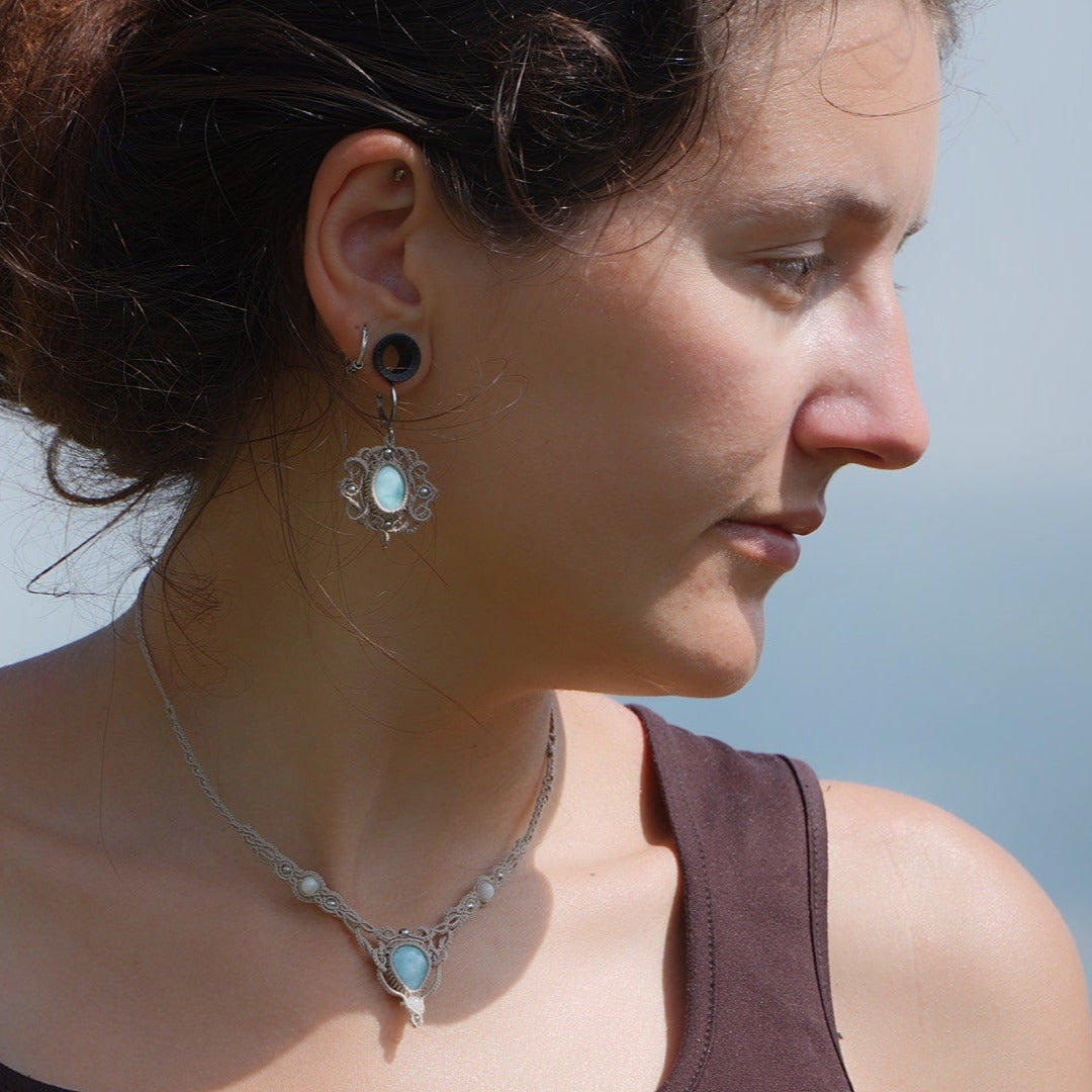 Parure Trésors d'Ecume composée d'une paire de boucles d'oreilles pendentes avec collier ras de cou assorti. Larimar bleu ciel ovales pour les boucles et en poire pointe en bas pour le collier. 3 Perles en nacre blanche pour le collier. Perles acier inoxydable argenté pour les deux pour le côté luxe et précieux. Dormeuses en acier inoxydable pour un port assuré. Fil de Coton couleur lin naturel (beige gris) de 0, 5 mm tissé en micro-macramé en relief version celtique. Bijoux porté femme brune peau caramel 