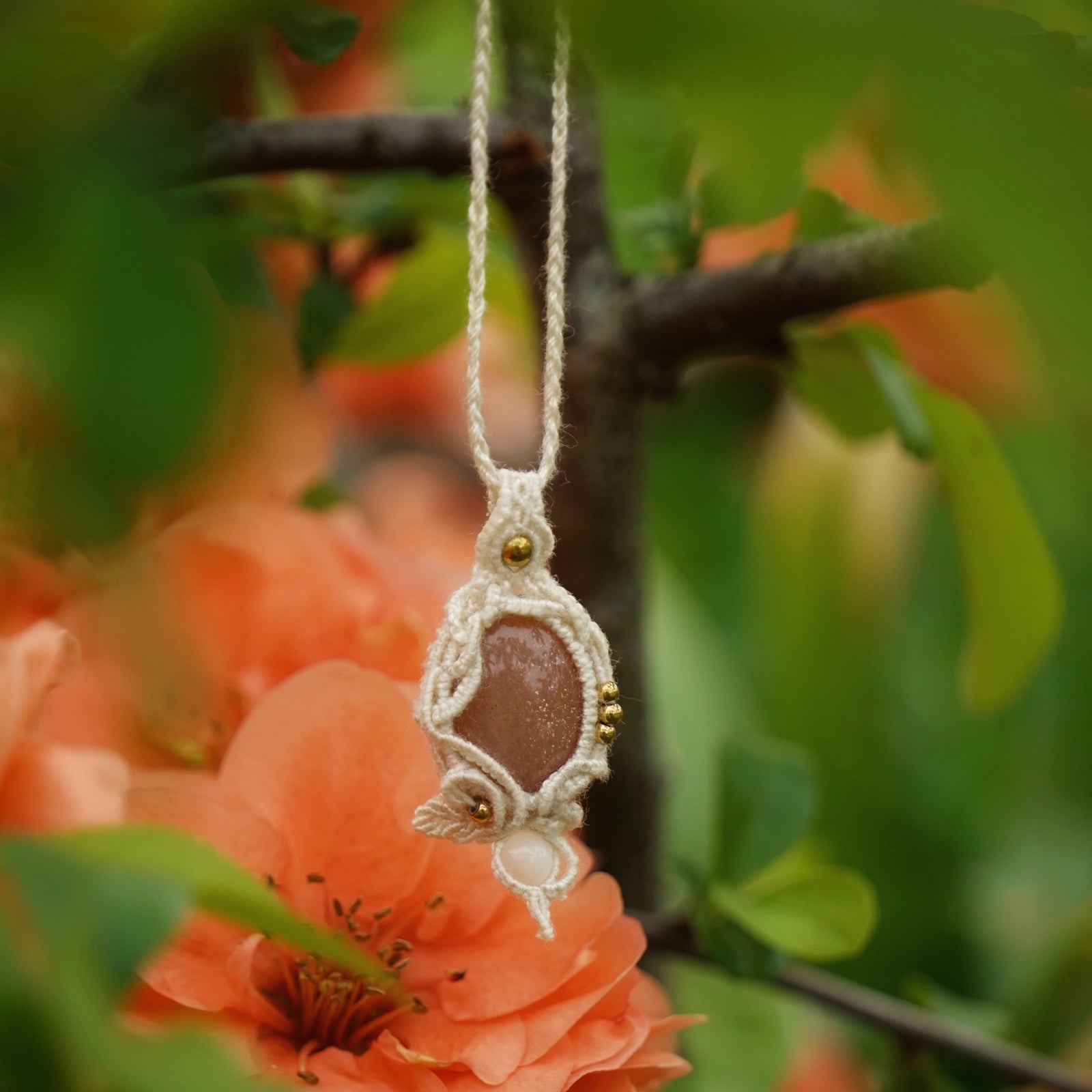 Collier pendentif en micro macramé avec pierre de soleil pêche et nacre, posé sur une branche de cognassier en fleurs saumonées, style bohème et féérique. Coton et Astrance