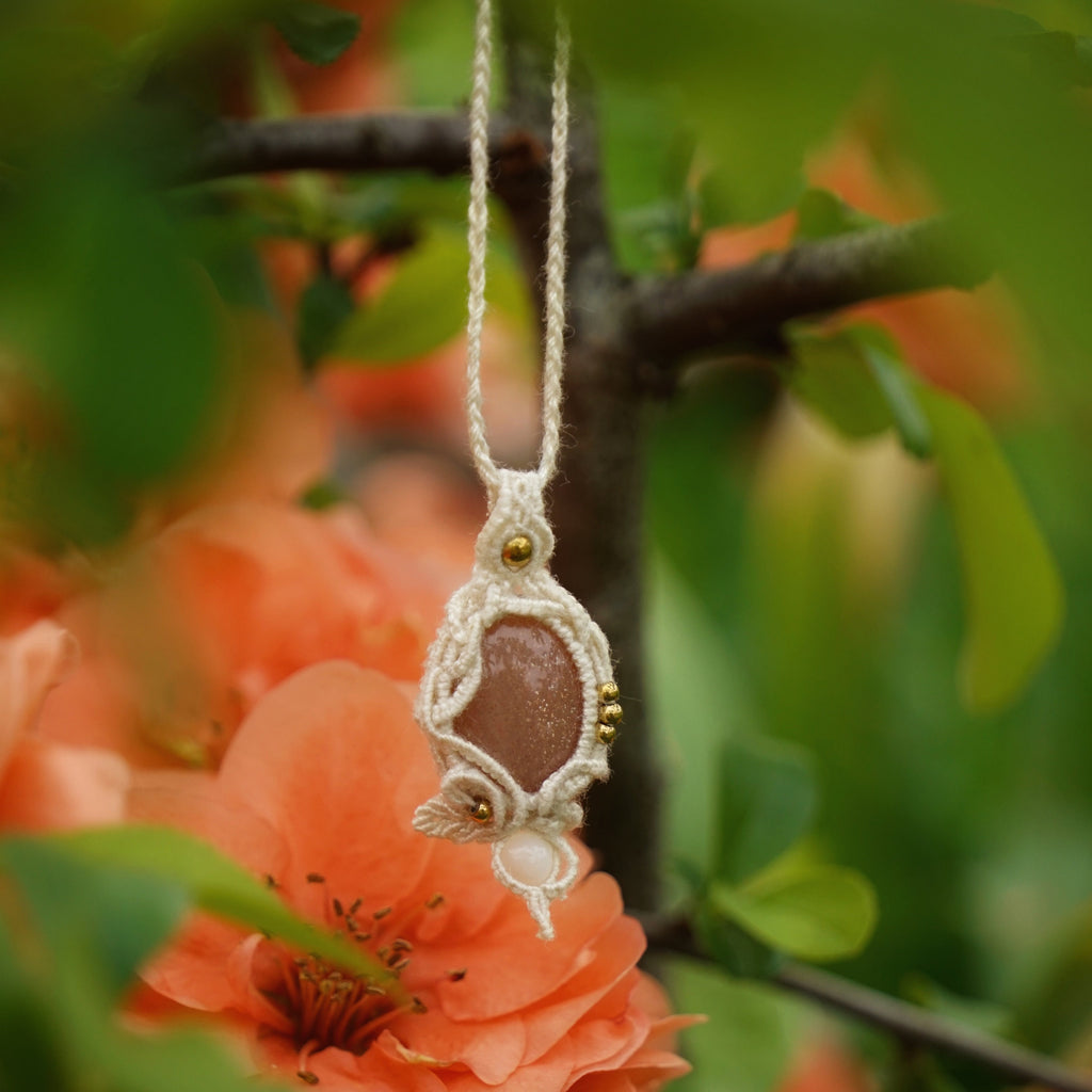 Collier pendentif en micro macramé avec pierre de soleil pêche et nacre, posé sur une branche de cognassier en fleurs saumonées, style bohème et féérique. Coton et Astrance