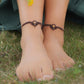 Bijoux de pieds, paire assortie de bracelets de chevilles à ambiance bohème celtique elfique. Fil marron foncé fin tissé en micro macramé avec entrelacs et arabesques, pierre centrale ovale petite taille jaune miel translucide. Bijoux hippie, bohème avec inspiration nature. Chevillères portées sur des chevilles à la peau pâle, bijoux de face, pieds posés dans de l'herbe avec à l'arrière une jupe vert forêt