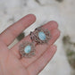 Boucles d'oreilles Trésors d'Ecume composée d'une Larimar bleu ciel ovales, mini Perles rondes en acier inoxydable argenté pour le côté luxe et précieux. Dormeuses en acier inoxydable pour un port assuré. Fil de Coton couleur lin naturel (beige gris) de 0, 5 mm tissé en micro-macramé en relief version celtique. Bijoux à plat dans une main à la peau claire avec fond en pierre blanche. Ambiance sirène, plage et tropiques, bohème et hippie. Parfait pour les tenues mariage et festivals. 