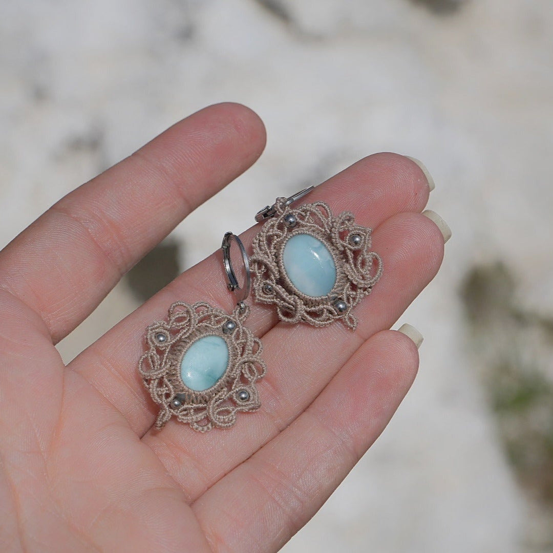 Boucles d'oreilles Trésors d'Ecume composée d'une Larimar bleu ciel ovales, mini Perles rondes en acier inoxydable argenté pour le côté luxe et précieux. Dormeuses en acier inoxydable pour un port assuré. Fil de Coton couleur lin naturel (beige gris) de 0, 5 mm tissé en micro-macramé en relief version celtique. Bijoux à plat dans une main à la peau claire avec fond en pierre blanche. Ambiance sirène, plage et tropiques, bohème et hippie. Parfait pour les tenues mariage et festivals. 