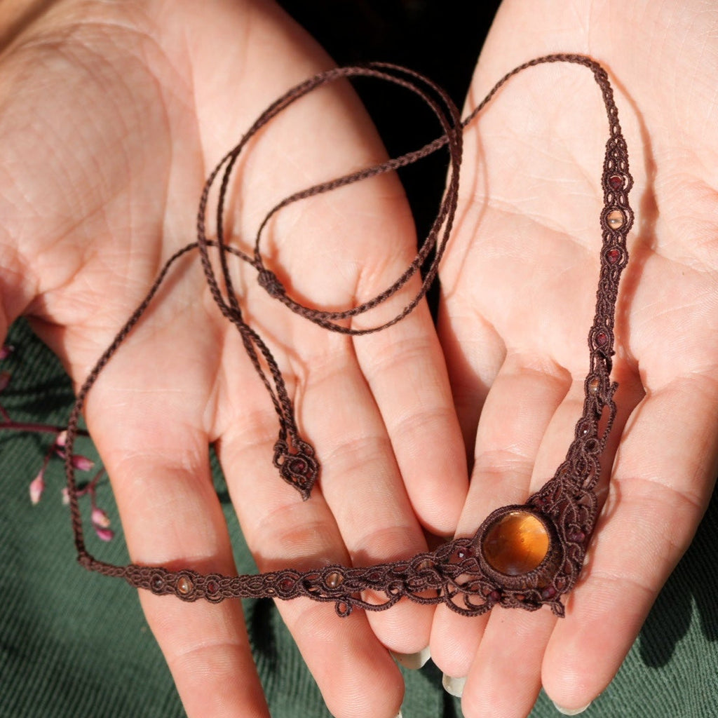 Bijou elfique artisanal réglable en citrine jaune et grenat rouge, tissé en macramé brun. Collier ras de cou bohème, présenté dans une main féminine à la peau claire, mettant en valeur la finesse du tissage.