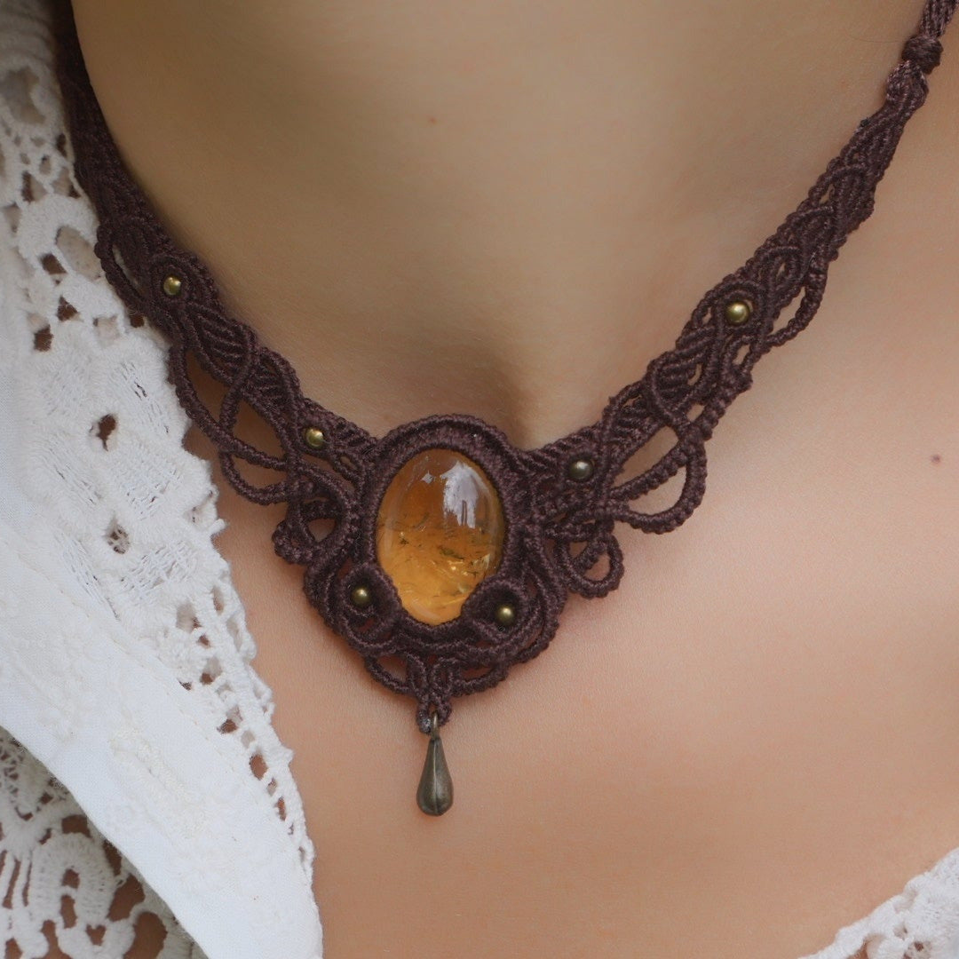 Collier large en micro macramé de couleur marron foncé, jaune ambré et bronze. Motif tissé en micro macramé avec ambiance entrelacs celtiques et arabesques elfiques. Présence d'une pierre centrale ovale de grande taille jaune translucide, gouttes bronze, forme V avec mini perles ronde bronze parsemées. Photo ambiance bohème hippie avec une touche d'élégance. Bijou porté sur une peau claire et chemise blanche en dentelle.