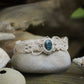 Bracelet large asymétrique. Bijou dentelle ambiance bohème hippie. Bracelet tissé en micro macramé avec fil de coton écru de 0.5 mm avec une pierre ovale de chrysocolle couleur bleu turquoise et marron foncé chocolat. Présence de part et d'autre de façon symétrique des perles en acier inoxydable argentées. Système de réglage à coulisse tubulaire à l'arrière. Design en relief et en torsade entrelacées celtiques. Ambiance plages, festival d'été et mariage boho. Bijou posé sur un galet beige et coquillages.
