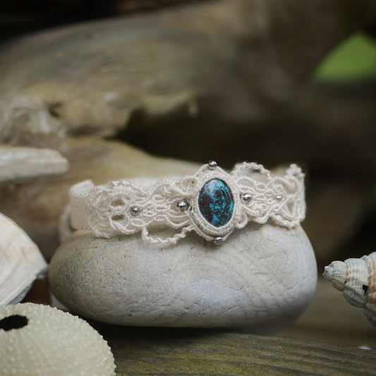 Bracelet large asymétrique. Bijou dentelle ambiance bohème hippie. Bracelet tissé en micro macramé avec fil de coton écru de 0.5 mm avec une pierre ovale de chrysocolle couleur bleu turquoise et marron foncé chocolat. Présence de part et d'autre de façon symétrique des perles en acier inoxydable argentées. Système de réglage à coulisse tubulaire à l'arrière. Design en relief et en torsade entrelacées celtiques. Ambiance plages, festival d'été et mariage boho. Bijou posé sur un galet beige et coquillages.