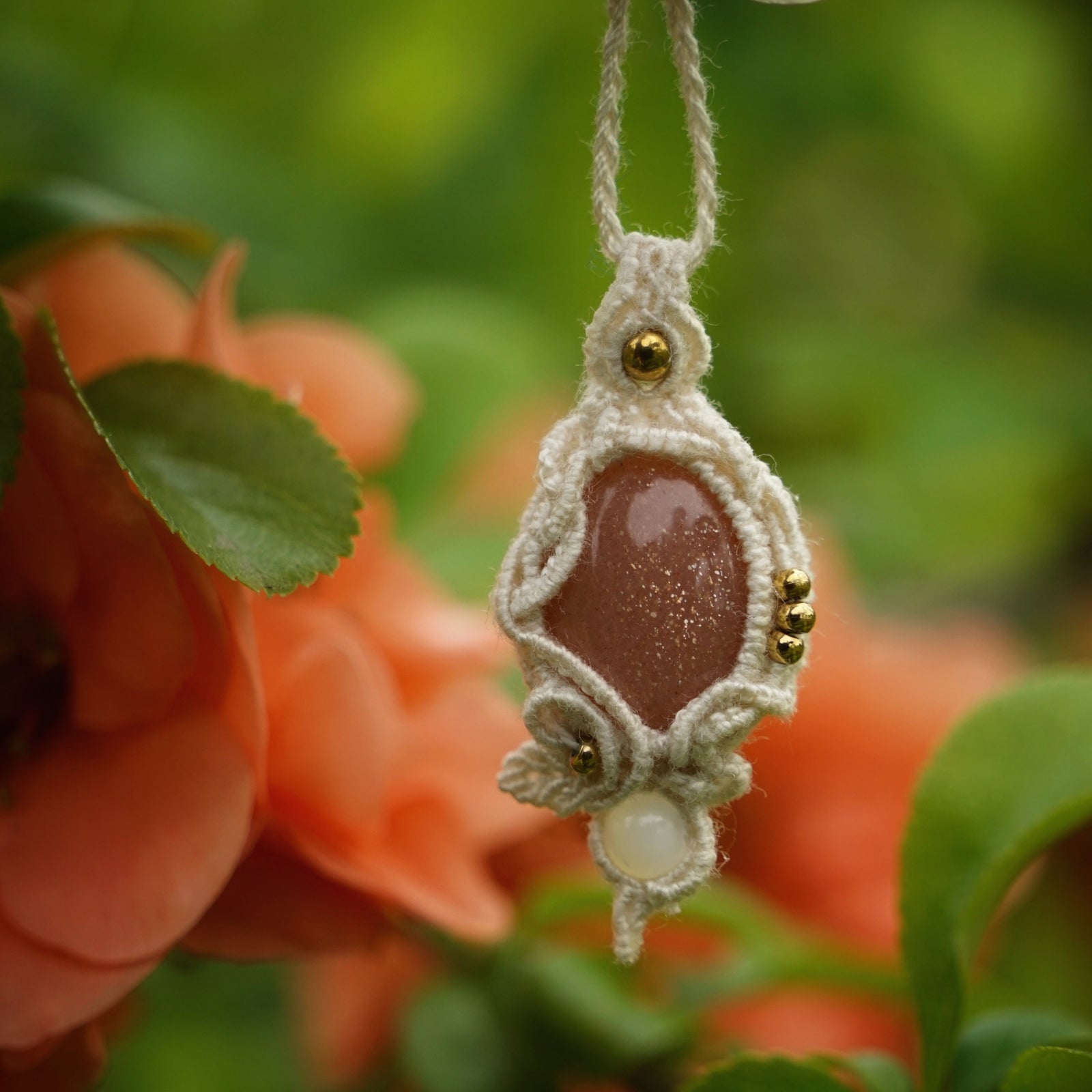 Collier pendentif en micro macramé avec pierre de soleil pêche et nacre, posé sur une branche de cognassier en fleurs saumonées, style bohème et féérique.