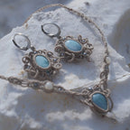 Parure Trésors d'Ecume composée d'une paire de boucles d'oreilles pendentes avec collier ras de cou assorti. Larimar bleu ciel ovales pour les boucles et en poire pointe en bas pour le collier. 3 Perles en nacre blanche pour le collier. Perles acier inoxydable argenté pour les deux pour le côté luxe et précieux. Dormeuses en acier inoxydable pour un port assuré. Fil de Coton couleur lin naturel (beige gris) de 0, 5 mm tissé en micro-macramé en relief version celtique. Bijoux posés sur une pierre blanche 