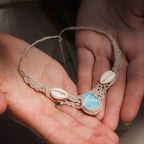 Collier plastron en Larimar Océan bleu ciel, coton couleur lin naturel (beige gris), cauris blancs et perles en acier inoxydable argenté. Système de réglable en tube tissé à l'arrière du bijou. Tissage symétrique en micro macramé avec effet nœuds celtiques entrelacés en relief. Style bohème, hippie, plage, Sirène, tribal et tropical. Convient pour un mariage type bohème ou plage. Bijou tenu vue 3/4, tenue romantique et bohème blanche avec dentelle apparente. Modèle à la peau bronzée et aux cheveux bruns. 