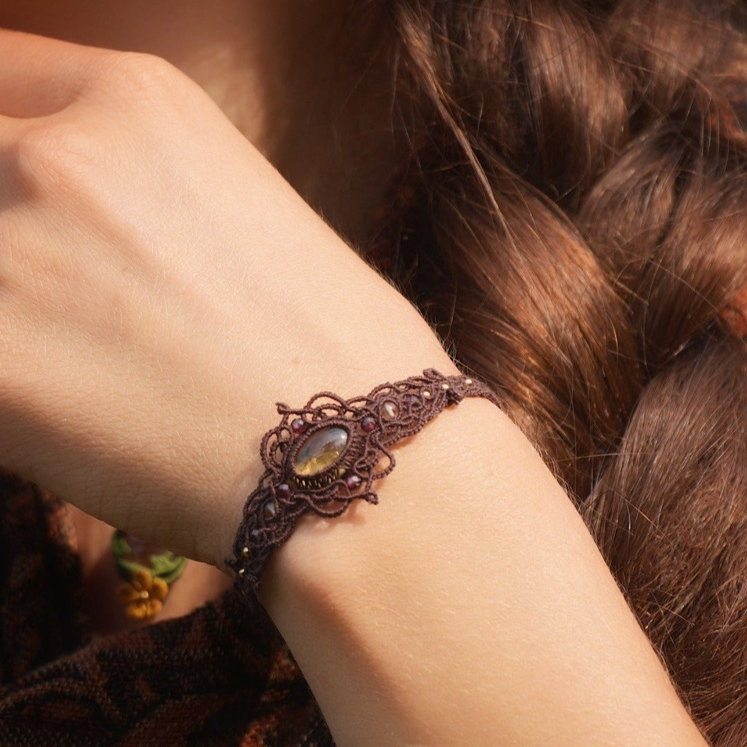 Bracelet large elfique en micro macramé brun, orné d’une citrine jaune pâle et de perles de grenat rouge rosé. Bijou bohème et artisanal réglable, porté au poignet d’une femme à la peau claire.